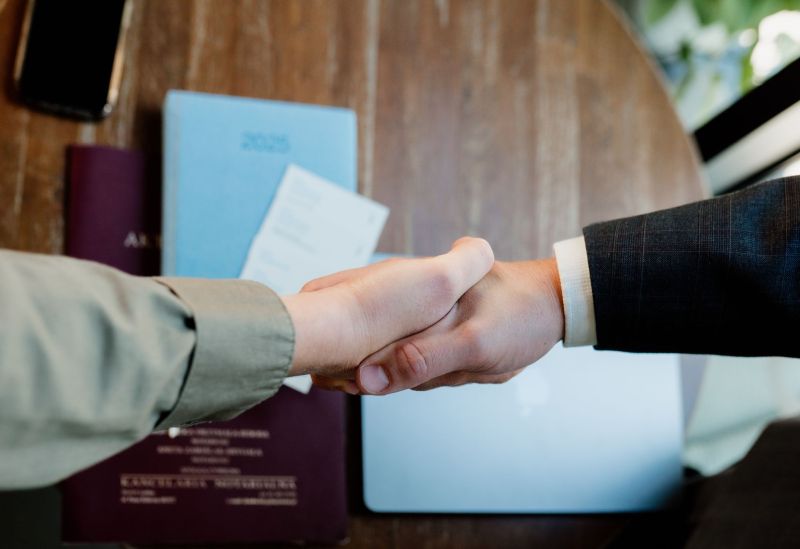 Uścisk dłoni przez kobietę i mężczyznę. W tle dokumenty i laptop lezące na stole