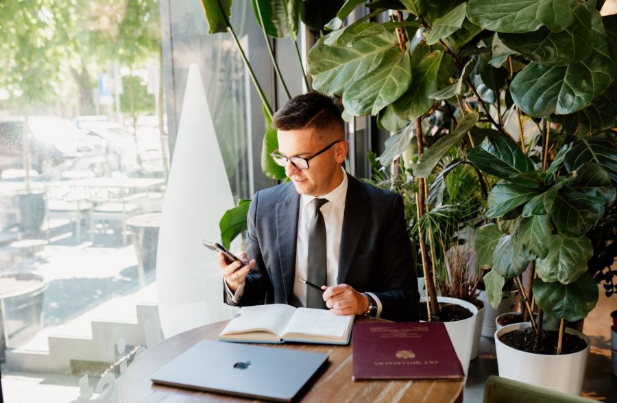Piotr Grudniewski siedzi przy stole i patrzy na telefon. Na stole leżą dokumenty. W tle widać kwiaty w dużych donicach.